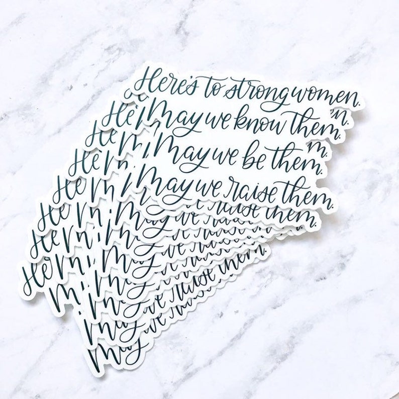 May include: A stack of white stickers with black lettering. The text on the stickers reads "Here's to strong women.  May we know them. May we be them. May we raise them."