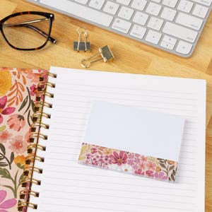 May include: A spiral-bound notebook with a floral cover featuring pink, orange, and yellow flowers, paired with a matching square notepad. The notepad has a white top section and a floral bottom section. Also includes glasses and two metal binder clips.
