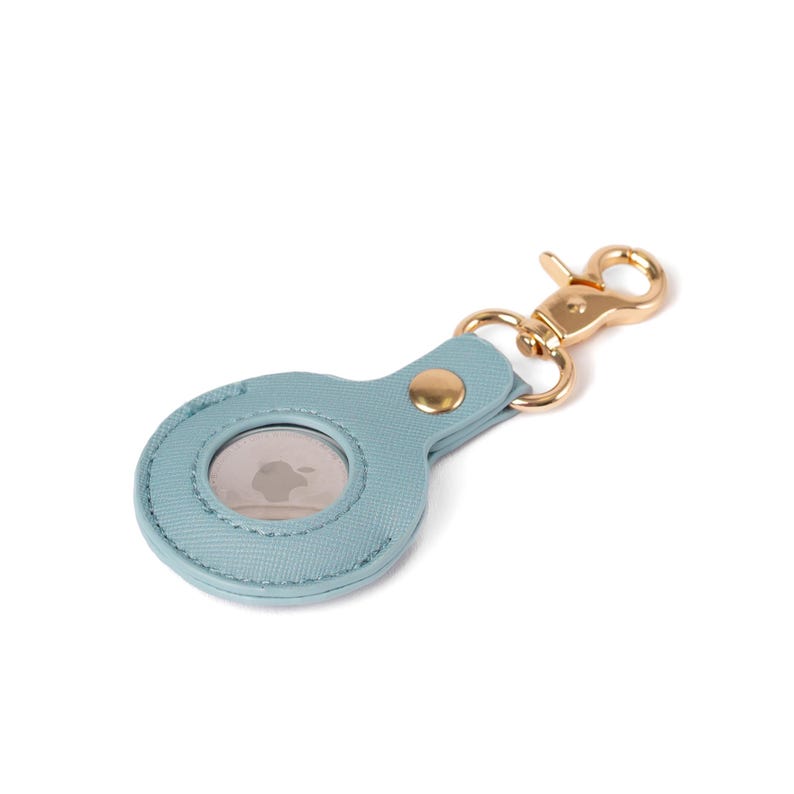 May include: A light blue textured protective holder for an Apple AirTag featuring a circular cutout to display the device. The holder includes a gold-toned metal snap button closure and a matching gold-toned lobster claw clasp for attachment.