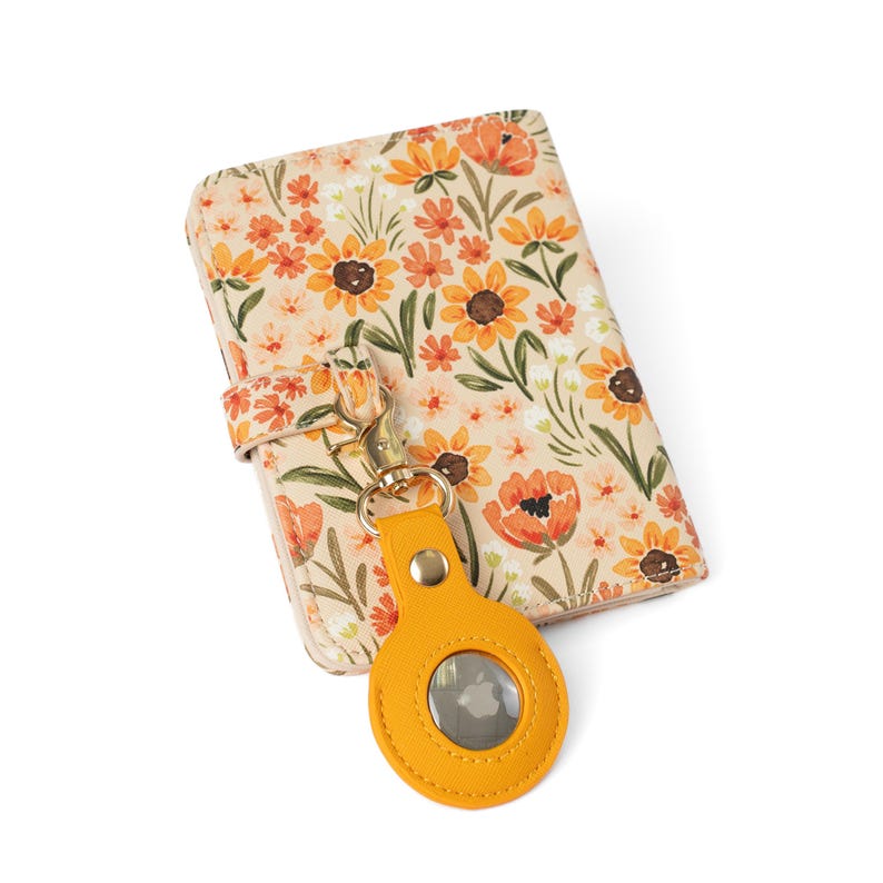 May include: A rectangular wallet featuring a cream background with a floral pattern of orange and yellow sunflowers and green leaves. A gold-toned metal clasp attaches a bright yellow circular leather-like tracker holder to the wallet exterior.