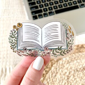 Puede incluir: Una pegatina transparente con una ilustraci&oacute;n de un libro abierto, rodeado por una corona floral. Las p&aacute;ginas del libro son blancas, con l&iacute;neas negras que representan el texto. La corona incluye hojas verdes, rosas amarillas y rosas y otros elementos florales.