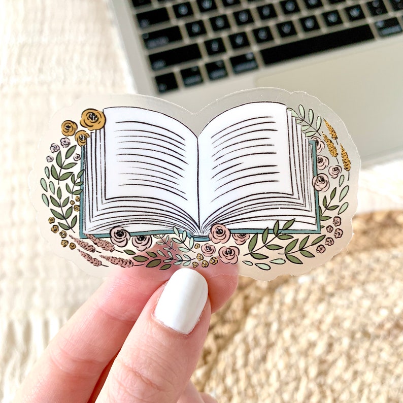 Puede incluir: Una pegatina transparente con una ilustraci&oacute;n de un libro abierto, rodeado por una corona floral. Las p&aacute;ginas del libro son blancas, con l&iacute;neas negras que representan el texto. La corona incluye hojas verdes, rosas amarillas y rosas y otros elementos florales.
