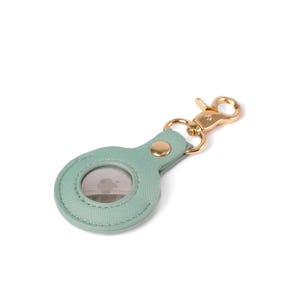 May include: A light mint green textured protective case for a circular tracking device. The case features a gold-toned metal lobster clasp attached by a matching green tab with a single gold-toned rivet, securing the device within the holder.