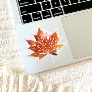 May include: A watercolor-style sticker of a brown maple leaf with a transparent background. The sticker is placed on a silver laptop.