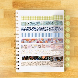 May include: A spiral notebook with nine rolls of decorative washi tape. The tapes feature various patterns, including gingham, floral, and stripes, in colors like green, blue, and pink. The tape rolls are arranged horizontally on the open notebook pages.
