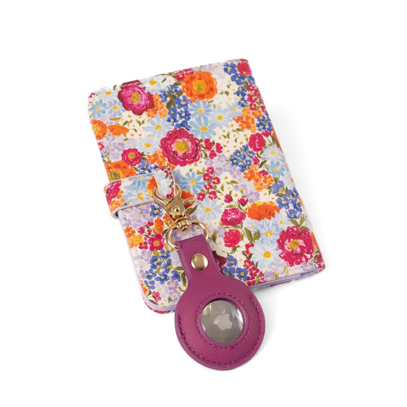 May include: A rectangular passport holder featuring a vibrant floral pattern with pink, orange, and blue flowers on a white background. A purple circular tracker case with a gold metal clasp is attached to the holder by a matching floral strap.