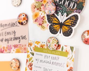 May include: A white magnetic board with a floral design. The board has a meal plan written on it with a black marker. The board is decorated with colorful floral magnets and a butterfly magnet. The text on the board reads "See the Good" and "Meal Plan".
