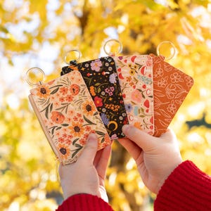 May include: Four floral-patterned cardholders with gold key rings. The cardholders feature different floral designs in shades of orange, black, and cream. The background is a blurred view of yellow leaves.