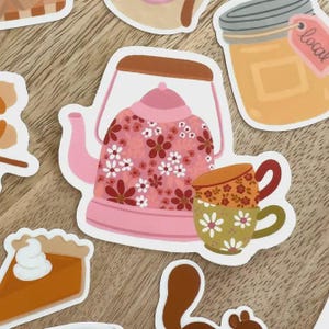 May include: A collection of stickers featuring a pink teapot with a floral pattern, two stacked teacups, a jar of honey with a "local" tag, and a slice of pumpkin pie. The stickers have a vintage, hand-drawn aesthetic.