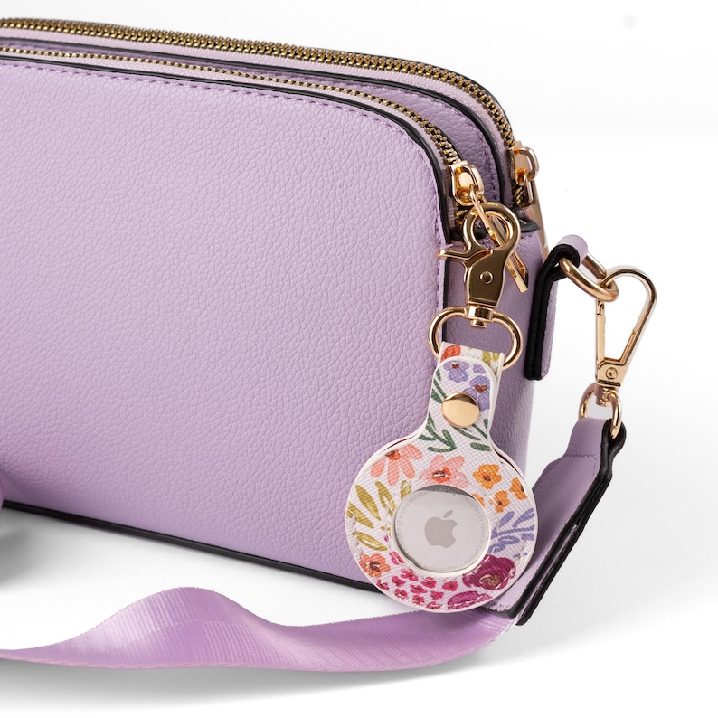 May include: A light purple crossbody bag with a gold zipper and hardware. A floral print AirTag holder is attached to the bag. The AirTag holder has a white background with colorful flowers.