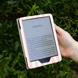 May include: A Kindle e-reader in a pink and white striped case, displaying text from "Chapter I" of a book. The e-reader is held against a backdrop of green foliage, showcasing the device's design and readability.