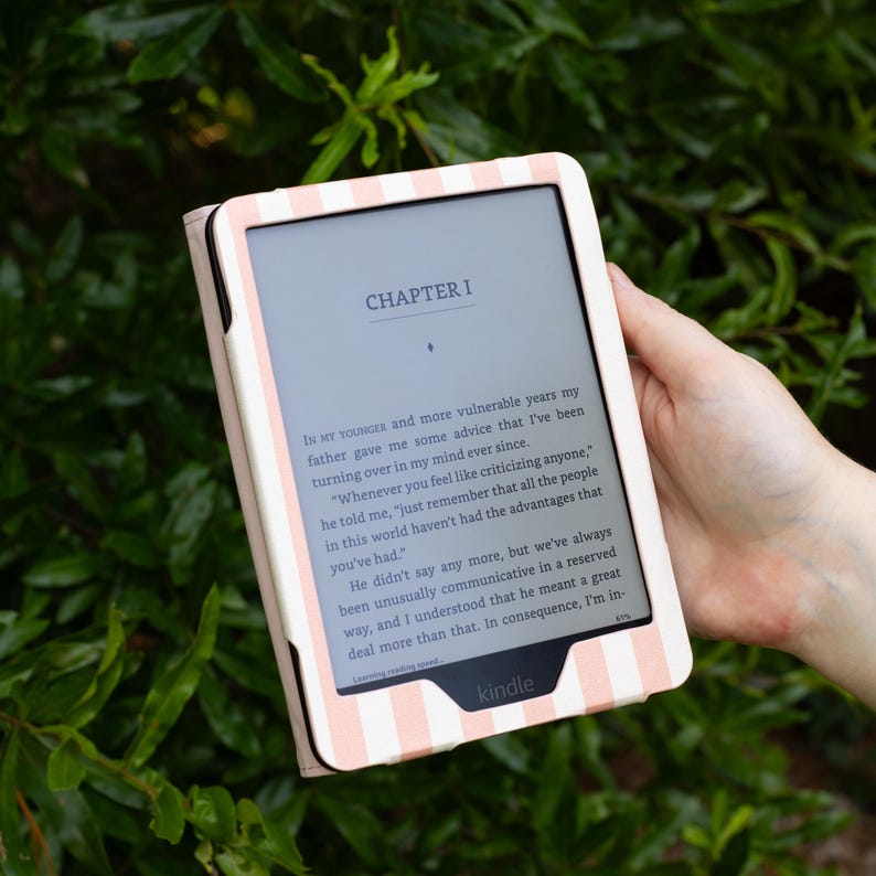 May include: A Kindle e-reader in a pink and white striped case, displaying text from "Chapter I" of a book. The e-reader is held against a backdrop of green foliage, showcasing the device's design and readability.