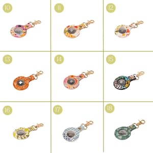 May include: A collection of AirTag holders in various floral and patterned designs. Each holder features a gold-colored clasp and a clear window to display the AirTag. The designs include pink, orange, yellow, blue, and black with floral and plaid patterns.