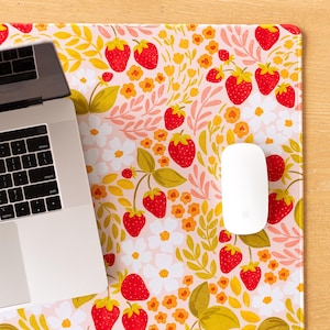 May include: A floral mousepad featuring a pattern of red strawberries, yellow flowers, and green leaves on a light pink background. A white computer mouse and a laptop are visible on the pad. The mousepad is designed for use with a computer.