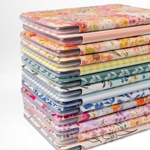 Può includere: Una pila di quaderni con motivi floreali e copertine di plastica trasparente. I quaderni presentano vari disegni, tra cui motivi floreali rosa, gialli e blu, e motivi a quadretti e a righe. I bordi sono arrotondati.