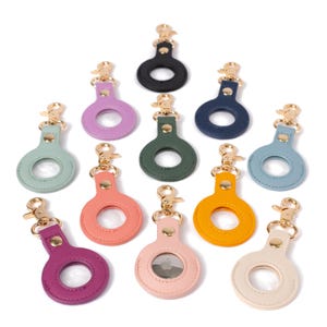 May include: A collection of eleven circular faux leather keychains in various colors, including black, navy, green, light blue, pink, yellow, and purple. Each keychain features a gold-toned metal lobster clasp and a central circular cutout.