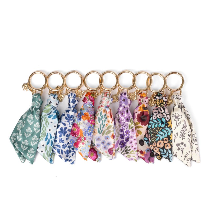 May include: A collection of ten keychains with colorful floral patterned fabric tassels. Each keychain has a gold-colored ring and a small charm. The fabric patterns include various flowers and leaves in shades of green, blue, purple, and orange.