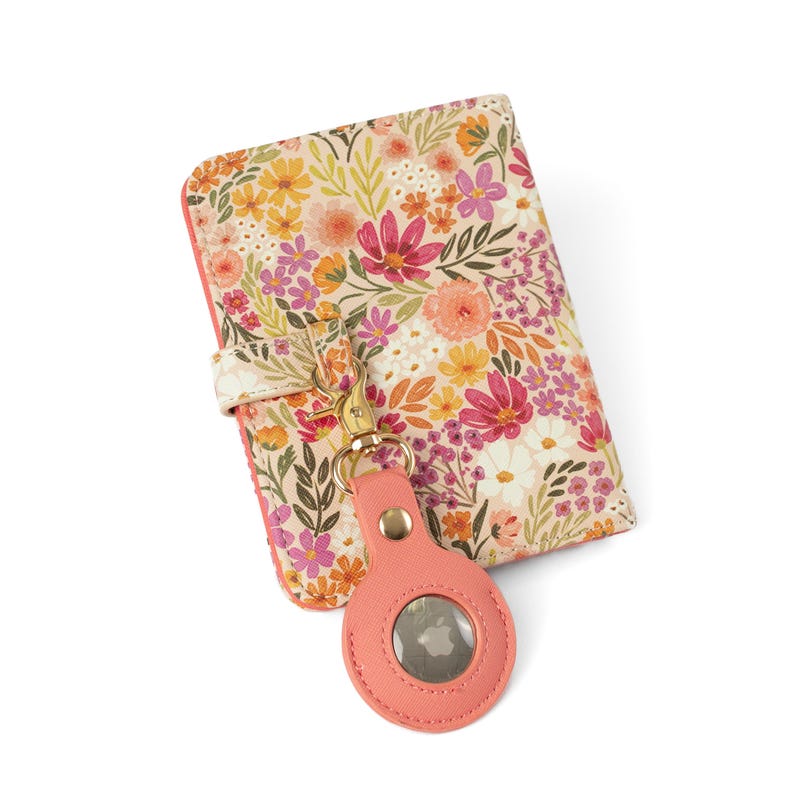 May include: A rectangular passport holder featuring a cream background with a colorful floral pattern in shades of pink, orange, yellow, and green. A coral-colored circular tracker case is attached to the holder by a gold-toned metal clasp.