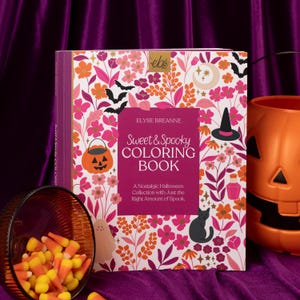 May include: A "Sweet & Spooky Coloring Book" with a floral and Halloween-themed cover. The book features a pumpkin, bats, and a witch's hat. Candy corn spills from a glass, and an orange jack-o'-lantern bucket is nearby.