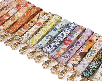 May include: A collection of floral patterned wrist lanyards with gold-colored lobster claw clasps. The lanyards feature various colors and designs, including orange, yellow, blue, and purple, with botanical and floral patterns. The lanyards are arranged in a diagonal row.