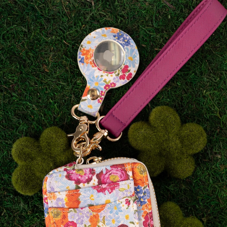 May include: Floral print AirTag holder with a matching wrist strap and a small wallet. The holder and wallet feature a colorful floral pattern with a gold-tone clasp. The wrist strap is a solid magenta color.