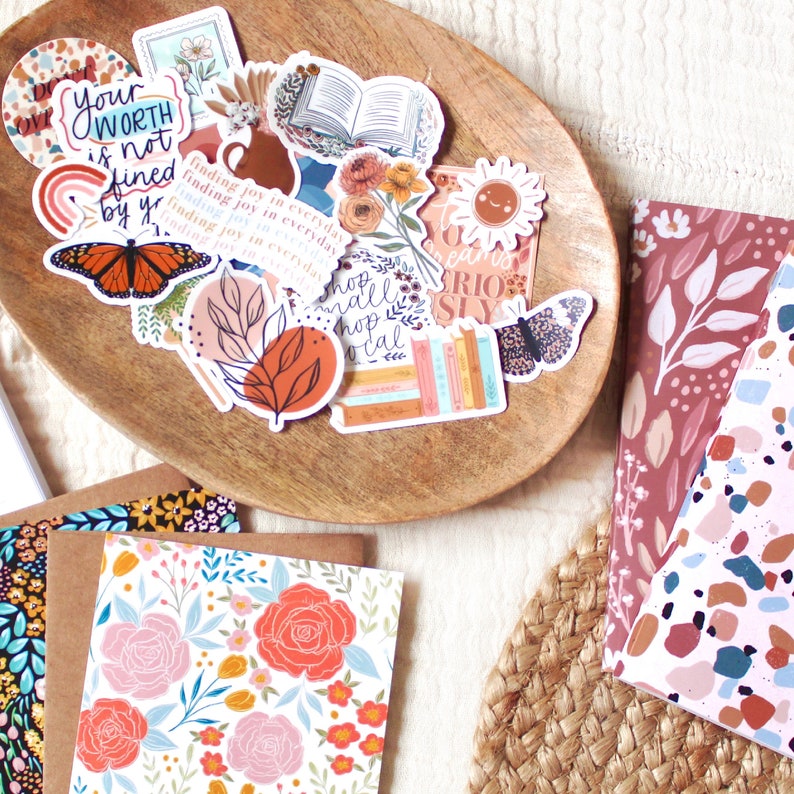 Puede incluir: Un cuenco de madera lleno de pegatinas coloridas con dise&ntilde;os florales, de mariposas e inspiradores. Tambi&eacute;n se ven tarjetas de felicitaci&oacute;n con motivos florales y cuadernos con dise&ntilde;os abstractos. Las pegatinas incluyen texto como "Your Worth is Not Fined by You."