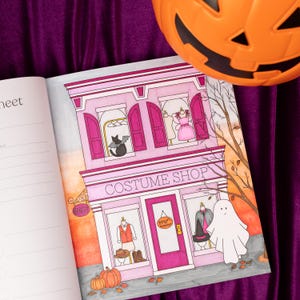 May include: A colorful illustration of a costume shop with a pink facade, featuring a black cat, dresses, and costumes in the windows. A ghost and pumpkins are also present. An orange plastic jack-o'-lantern sits in the upper right corner.