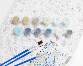May include: A paint-by-numbers kit with a floral design. The kit includes numbered paint pots, paintbrushes, and a color guide. The canvas features a detailed floral pattern in shades of blue and gray. The paint pots are arranged in a row, numbered 1 through 12.