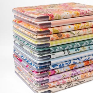 May include: A stack of notebooks with floral and patterned covers. The covers feature various designs, including pink, yellow, and blue floral patterns, and gingham and striped patterns. The notebooks have clear plastic covers.