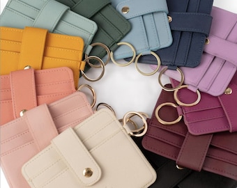 Pode incluir: Uma coleção de porta-cartões coloridos em várias tonalidades, incluindo amarelo, rosa, azul e preto. Cada porta-cartões apresenta um fecho de pressão e um porta-chaves dourado. O nome da marca "elyse trebonne" é visível.