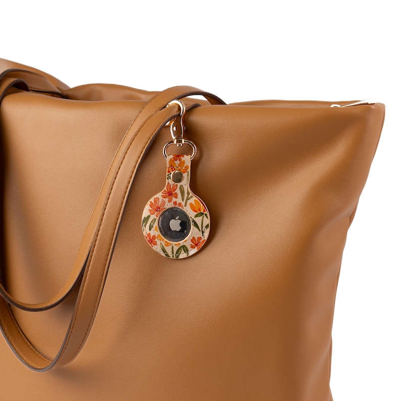 May include: A floral-patterned AirTag holder with a gold-colored clasp is attached to a tan leather tote bag. The holder features a design of orange and red flowers on a cream background. The tote bag has a zipper closure and a shoulder strap.