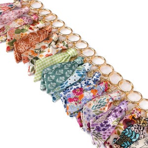 May include: A collection of floral patterned keychains with gold-colored rings. The fabric patterns include various colors such as pink, orange, green, blue, and purple. The keychains are arranged in a diagonal line against a white background.
