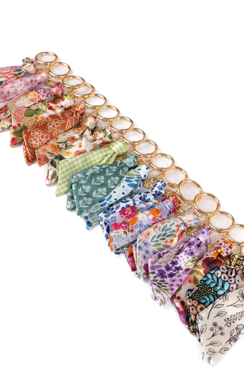 May include: A collection of floral patterned keychains with gold-colored rings. The fabric patterns include various colors such as pink, orange, green, blue, and purple. The keychains are arranged in a diagonal line against a white background.