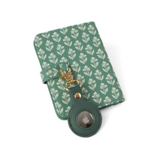 May include: A sage green passport holder featuring a repeating white floral pattern, secured with a matching strap. A gold-toned metal lobster clasp connects the holder to a circular, sage green protective case containing a silver Apple AirTag.