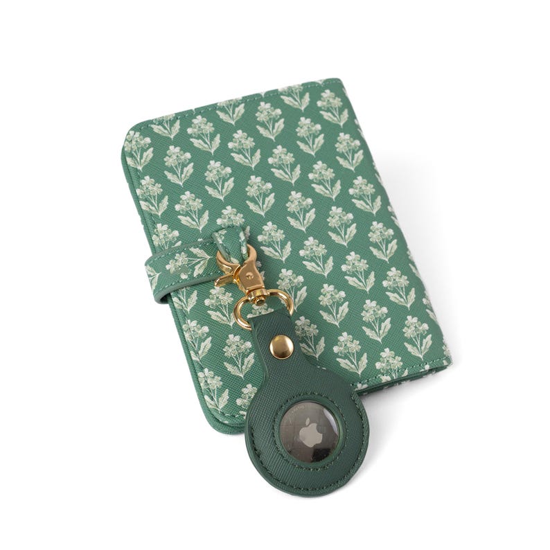 May include: A sage green passport holder featuring a repeating white floral pattern, secured with a matching strap. A gold-toned metal lobster clasp connects the holder to a circular, sage green protective case containing a silver Apple AirTag.