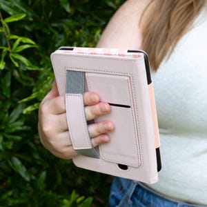 May include: A light pink tablet case with a striped pattern at the top. The case has a hand strap and a gray elastic band for secure handling. The case is held in front of a green leafy background.