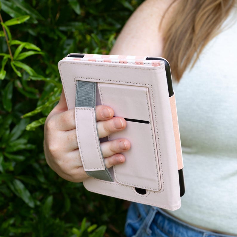 May include: A light pink tablet case with a striped pattern at the top. The case has a hand strap and a gray elastic band for secure handling. The case is held in front of a green leafy background.
