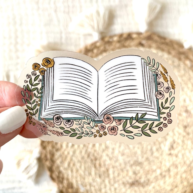 Puede incluir: Una pegatina con una ilustraci&oacute;n de un libro abierto con l&iacute;neas negras. El libro est&aacute; rodeado por un borde floral de hojas verdes, rosas rosas y amarillas y ramitas de lavanda. La pegatina tiene un fondo transparente.