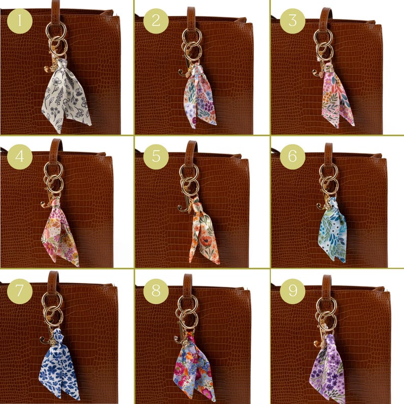 May include: Nine different floral patterned silk scarf keychains attached to a brown leather bag. Each keychain has a gold-colored ring and hook. The scarves feature various colors and designs, including white, blue, pink, and purple.