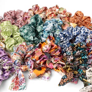 May include: A collection of colorful satin scrunchies in various floral and patterned designs. The scrunchies feature shades of pink, orange, blue, green, and purple. Some have attached bows. These hair accessories are perfect for adding a touch of style.
