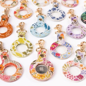 May include: A collection of floral-patterned AirTag holders with gold-colored clasps. The holders feature various designs, including orange, yellow, pink, blue, and purple floral prints. Each holder has a circular opening to display the AirTag.