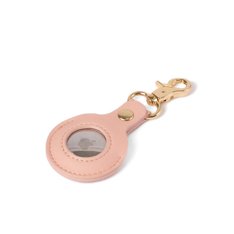 May include: A light pink textured faux leather protective case for a circular tracking device. The case features a gold-toned metal lobster clasp attached by a matching pink tab with a single gold-toned metal snap button closure.