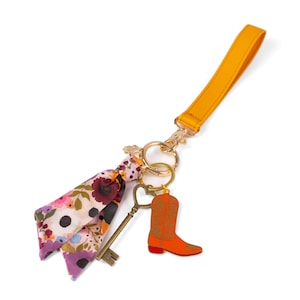 May include: A yellow leather wrist strap with gold-colored hardware, attached to a keychain with a floral scarf, a key, and an orange cowboy boot charm. The boot charm has green and orange details.