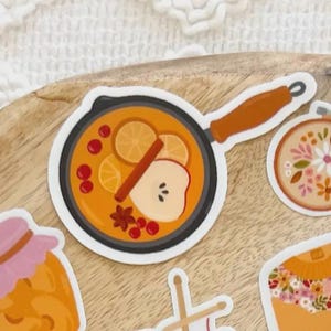 May include: A sticker featuring a cartoon illustration of a small pan filled with a warm beverage. The beverage includes orange slices, a cinnamon stick, an apple slice, and berries. The pan has a black base and a brown handle.