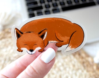 May include: A clear sticker with a cartoon illustration of a sleeping fox. The fox is curled up with its tail wrapped around its body. The fox is brown with a white belly and a black nose.