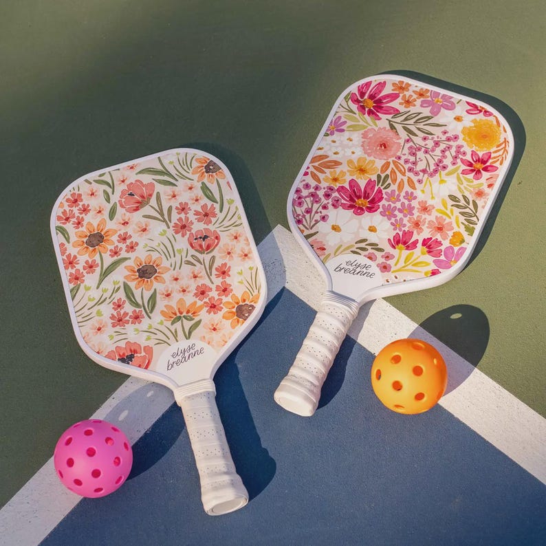 May include: Two pickleball paddles with floral designs in shades of pink, orange, and yellow. The paddles have white handles and are next to two pickleballs, one pink and one orange. The paddles have the text "elyse breanne".