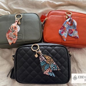 May include: Three crossbody bags in olive green, orange, and black. Each bag has a gold-tone ring with a floral scarf and a tassel. The black bag has a quilted pattern. The bags are on a neutral background.