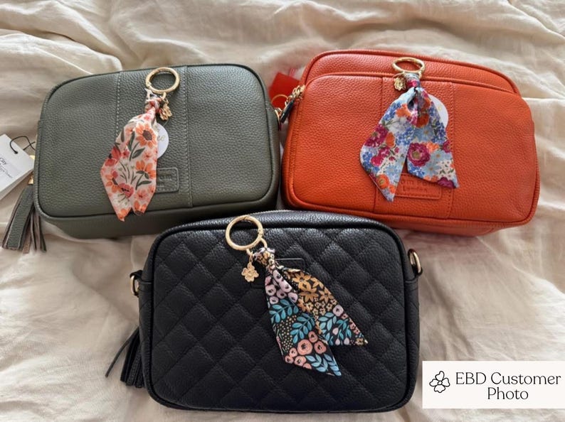 May include: Three crossbody bags in olive green, orange, and black. Each bag has a gold-tone ring with a floral scarf and a tassel. The black bag has a quilted pattern. The bags are on a neutral background.