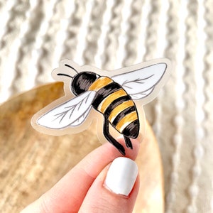 May include: A clear sticker with a cartoon illustration of a bee with black and yellow stripes. The bee is flying with its wings spread out.