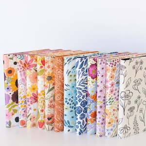 May include: A collection of floral-patterned journals in various colors. The journals feature designs with sunflowers, roses, and other flowers. The covers are in shades of orange, pink, blue, and white. The journals are standing upright.
